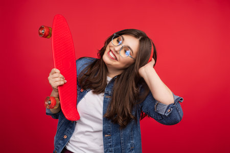 Smiling Woman Holding Red Skateboard Against Vibrant Red Backgroundの写真素材