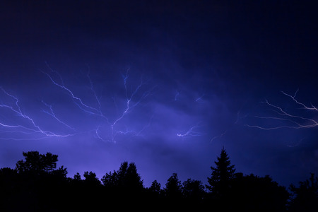 Lightning arcs through the summer night sky over silhouetted trees in the Midwestern United States 