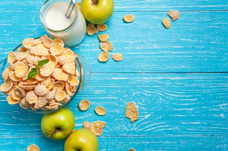 Bowl of cornflakes cereal on a blue wooden table and fresh apple, milk behind. Horizontal image with...
