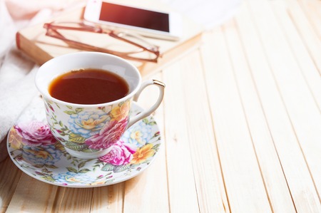 Tea cup placed on a wooden table with a smart phone, glasses, books and light cozy cloth. Copy space. Toned.の写真素材