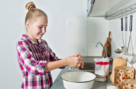 Young beautiful smiling girl breaks an egg into deep dish. Cute blonde cooking in a home kitchenの写真素材