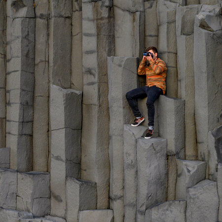 Man sitting on basalt rock taking picture with camera, Reynisfjara Black Sand Beach, Vik, Myrdalshreppur, Southern Region, Icelandのeditorial素材