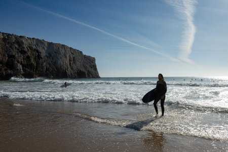 Surfer holding a surf board walking along the beach coast of Sagres that sits at the extreme western...