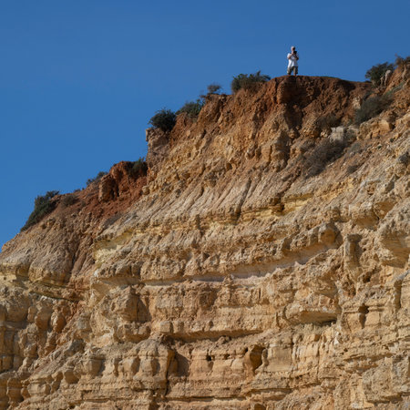 Tourist standing on the ledge of a cliff at The Praia do Porto de Mos that is the second largest beach in the Lagos region of Portugalのeditorial素材