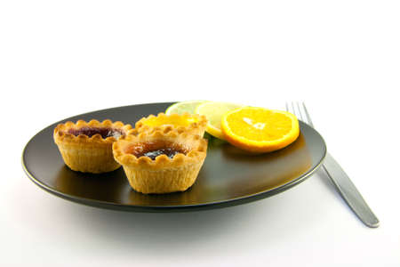 Red and yellow small jam tarts with slices of lemon, lime, and orange on a black plate with a fork on a white backgroundの写真素材