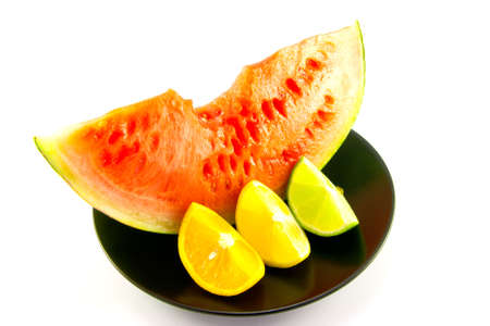 Slice of red juicy watermelon with a wedge of lemon, lime and orange on a black plate with a white backgroundの写真素材
