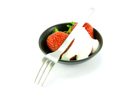 Sliced and whole red fresh strawberries with cream and small fork in a small black dish on a white backgroundの写真素材