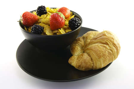 Cornflakes with strawberries and blackberries in a round black bowl with a croissant on a white backgroundの写真素材