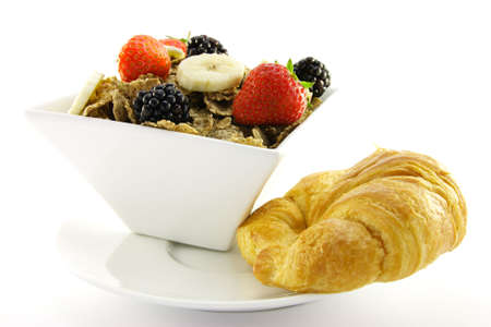 Crunchy looking delicious bran flakes and juicy fruit in a white bowl with a croissant on a white backgroundの写真素材