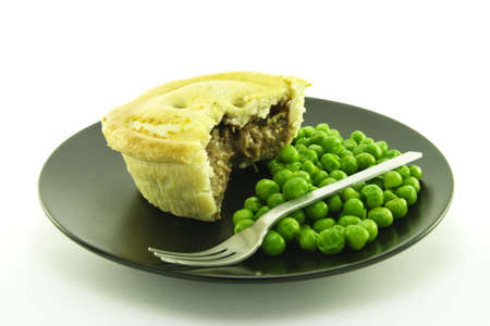 Small round cooked meat pie with green peas on a black plate with a small fork on a white background