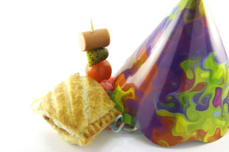 Small sausage roll with cone party hat and cocktail stick containing hot dog sausage, gherkin and tomato on a reflective white backgroundの写真素材