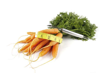 Fresh young bunch of carrots with green leafy stems and tape measure and chrome peeler on a reflective white backgroundの写真素材