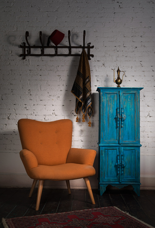 Composition of vintage orange armchair, blue cupboard, wall hanger with ornate scarf and red fez in studioの写真素材