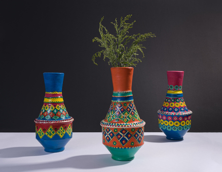 Still life of three artistic painted colorful handcrafted pottery vases and little green branches with harsh shadow on white table and black wallの写真素材