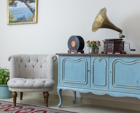 Vintage interior of retro off white armchair, vintage wooden light blue sideboard, old phonograph (gramophone) and vinyl records on background of beige wall, tiled porcelain floor, and red carpetの写真素材