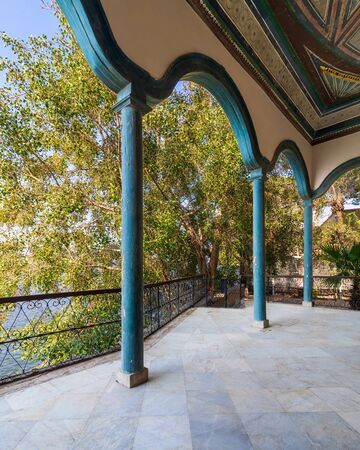 Covered terrace with green stone pillars, and marble floor fenced with metal forged fence and surrounded with green trees in sunny summer dayの写真素材