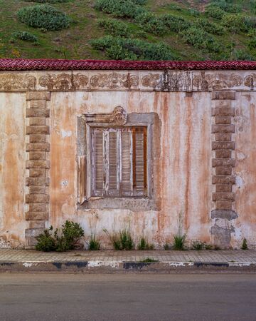 Exterior of aged house with weathered ornamental walls and shuttered window located under green hill...
