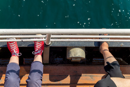 Top view showing legs of two persons, sitting on a narrow sea side passage of a ferry boat, looking toward the sea, and putting their legs on passage handrailの写真素材