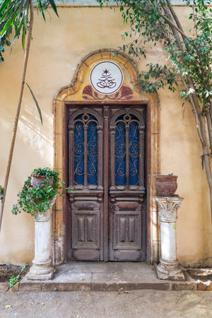 A closeup of Grungy traditional Egyptian style intricately carved wooden door, with wrought iron decorations in a yellow stone wall surrounded by bushes in flower potsの写真素材