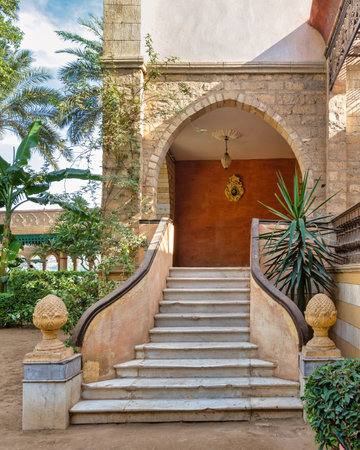 White marble staircase with ornate stone railings, flanked by sculptures, that descends to a serene courtyard surrounded by lush greenery and tropical plantsの写真素材