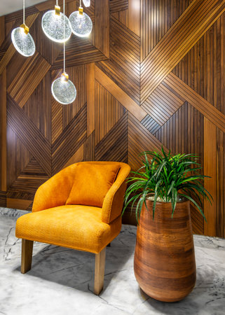 Modern orange armchair, beside tall wooden planter with green bushes, and contemporary tall glass chandelier, in a hall with decorated wood cladding wall, and white marble floorの写真素材