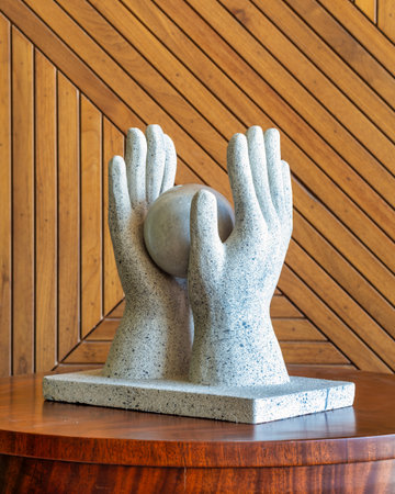 A stone sculpture depicts two hands gently cradling a round sphere, set on a wooden table with a...