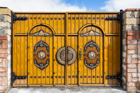 A beautifully crafted wooden gate stands prominently against a backdrop of stone walls. The gate features elaborate decorations and shiny metallic handles, illuminated by bright sunlight.の写真素材