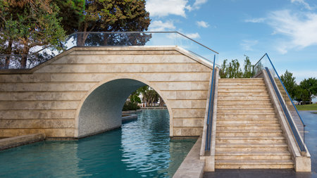 The Boulevard canals, or Little Venice, Baku, Azerbaijan: Stone bridge with arched passage over a clear blue water canal, adjacent to stairs with glass railings, surrounded by greenery under blue skyの写真素材