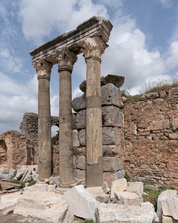 Historic ruins featuring three ancient columns with ornate capitals and stone walls at Ephesus,...