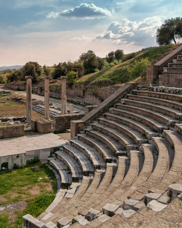 Historic Roman Theatre at Asclepieion of Pergamon, Bergama, Turkey. Ancient tiered seating and columns amidst green hills under a dramatic sky.の写真素材