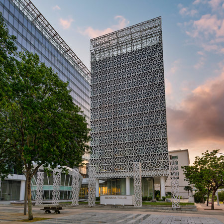Menara Tulus, a modern building with a geometric facade, alongside a glass structure and trees at...