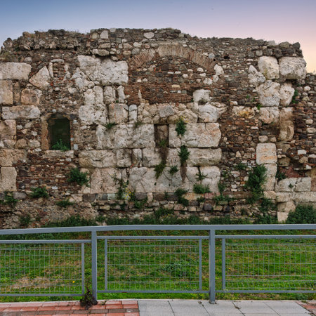 Agora of Smyrna, Izmir, Turkey. Ancient Roman ruins: weathered stone and brick wall with arch, openings, and vegetation. Metal fence, green grass, and soft sky.の写真素材