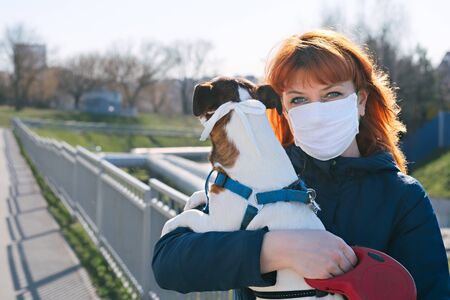 A red-haired girl in the backlight in a protective mask with a dog in her arms, because masks are required outside the house during a coronavirus infection.の写真素材