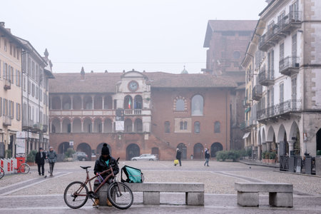Migrant sits alone on a bench in a square in the center of Pavia with a bicycle and looks at the phone: Pavia, Italy, Lombardy - January 28, 2020のeditorial素材