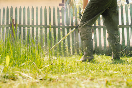 Man mows tall grass with an electric trimmer in the yard of country house. Mowing the lawn with hand mowerの写真素材