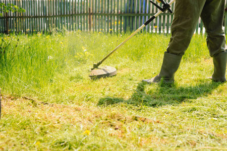 Man mows tall grass with an electric trimmer in the yard of country house. Mowing the lawn with hand...