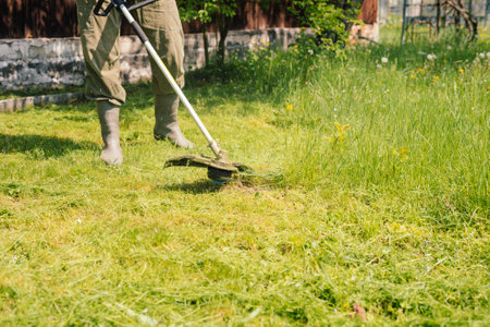 Man mows tall grass with an electric trimmer in the yard of country house. Mowing the lawn with hand...