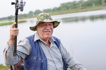 Happy mustachioed positive cheerful old man enjoying fish, hold a fishing rod, sit on a chair. Leisure and outdoor hobbiesの写真素材
