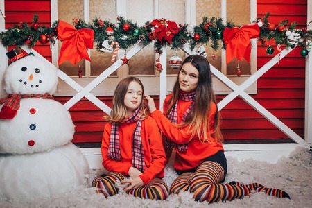 Two girls are sisters in red sweaters and striped scarves relax and have fun in a beautiful room, decorated for Christmas and the New yearの写真素材