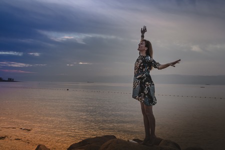 Girl on the rocks above sea and evening sky with sunset and blue clouds behindの写真素材