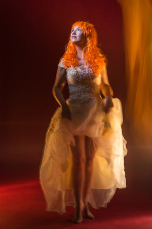 Old female actress with orange hair dancing in white dress with light show with yellow, orange and green lights around and red backgroundの写真素材
