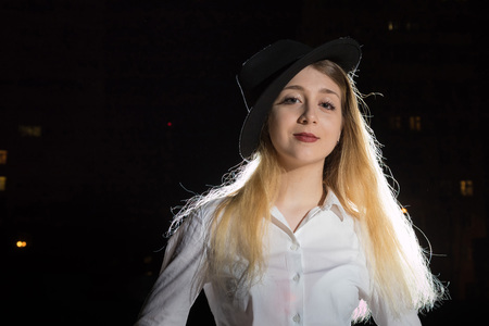 Girl in white shirt and black hat outdoors in a night and light behind herの写真素材