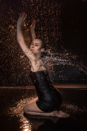 Girl in black dress in water in a small pool, stream of water flying to her and black background during a photoshoot with waterの写真素材