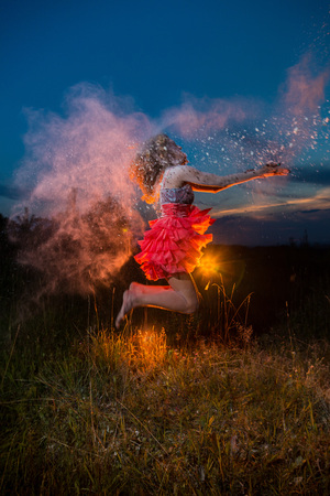 A girl in a black top and red skirt jumping up in a field and a dust cloud behind her. Model during a photo shoot with flour at sunsetの写真素材
