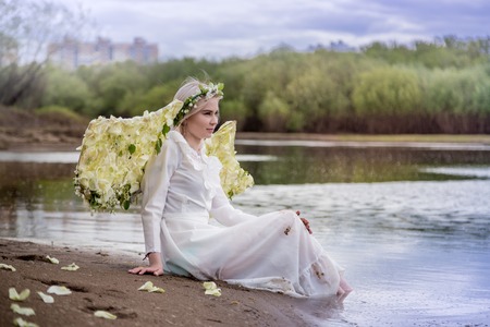 Nice blonde girl with white flower wings on the beach near river in cloudy dayの写真素材