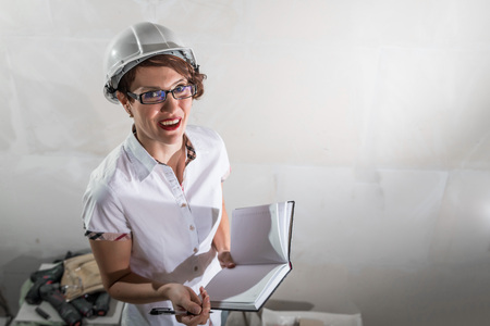 Cute nice young woman in white construction protection building helmet and unfinished apartment in the background.の写真素材