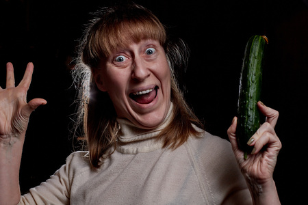 Middle-aged woman holding an cucumber in her hand and she does not like it. Denial of a healthy lifestyle. Photoshoot in dark keyの写真素材