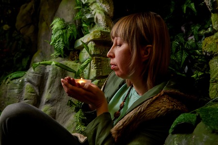 Girl in a green jacket with blond hair with a small candle in her hands near an artificial rock with...
