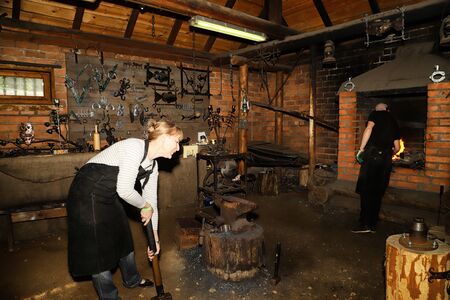 Dudutki, Belarus - September 13, 2018: Blacksmith working on an anvil together with female tourist...