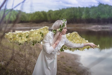 Nice blonde girl with white flower wings on the beach near river in cloudy dayの写真素材